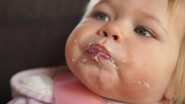 Happy cute chubby baby girl open mouth very wide waiting for food eating from spoon close up face covered in milk porridge food
