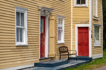 Two red doors on a yellow exterior wall of an old house. The building has multiple antique double hung windows with white trim and lace curtains. A black metal bench sits on a grey wooden step.