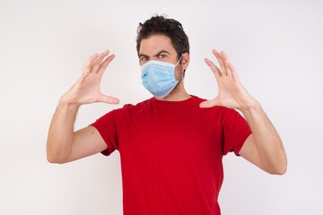 Young caucasian man with short hair wearing medical mask standing over isolated white background Shouting frustrated with rage, hands trying to strangle, yelling mad.