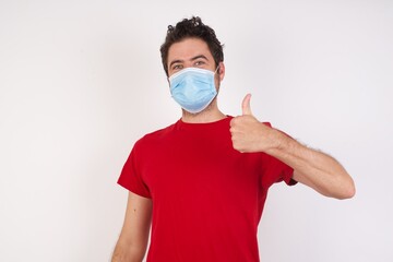 Young caucasian man with short hair wearing medical mask standing over isolated white background doing happy thumbs up gesture with hand. Approving expression looking at the camera showing success.