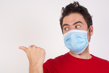 charming Young caucasian man with short hair wearing medical mask standing over isolated white background looking aside and indicaring finger side empty space