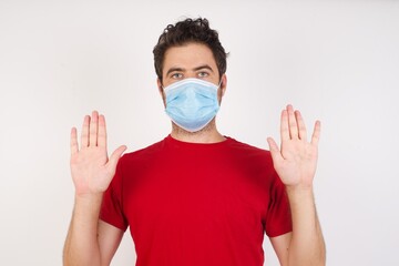 Serious Young caucasian man with short hair wearing medical mask standing over isolated white wall pulls palms towards camera, makes stop gesture, asks to control your emotions and not be nervous
