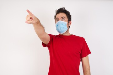 Young caucasian man with short hair wearing medical mask standing over isolated white background Pointing with finger surprised ahead, open mouth amazed expression, something on the front.