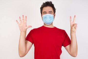 Young caucasian man with short hair wearing medical mask standing over isolated white background showing and pointing up with fingers number seven while smiling confident and happy.