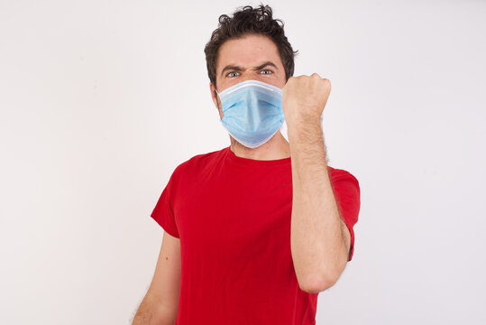 Young Caucasian Man With Short Hair Wearing Medical Mask Standing Over White Wall Being Angry And Mad Raising Fist Frustrated And Furious While Shouting With Anger. Rage And Aggressive Concept.