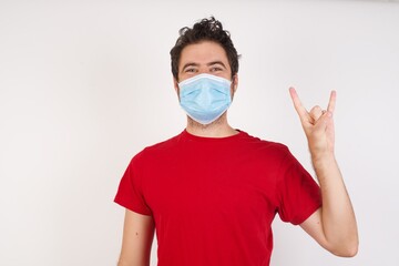 Young caucasian man with short hair wearing medical mask standing over isolated white background  doing a rock gesture and smiling to the camera. Ready to go to her favorite band concert.