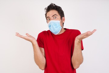 Frustrated Young caucasian man with short hair wearing medical mask standing over isol feels puzzled and hesitant, shrugs shoulders in bewilderment, keeps mouth widely opened, doesn't know what to do.