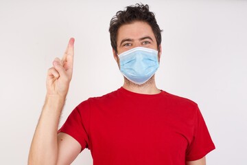 Young caucasian man with short hair wearing medical mask standing over isolated white background pointing up with fingers number ten in Chinese sign language Shi