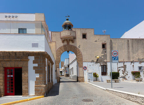 White buildings at the port of the Spanish city of Rota. In the middle is an archway with an old beacon and a narrow alley with a tower at the end.