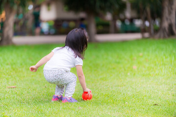 Asian girl plays a small orange ball on green lawn in park. Cute kid in white was pick up down on...