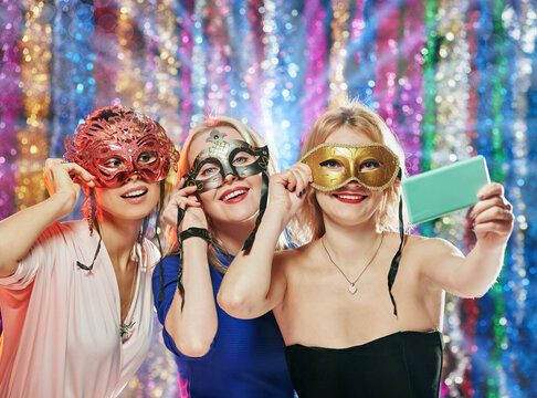 Three happy excited girls in carnival masks photographing on xmas party