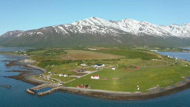 Aerial view around a small village on the coast of the Barents sea, snowy mountains in the background, sunny, summer day, in Troms, Nordland, North Norway -  orbit, drone shot