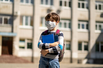 COVID-19 coronavirus quarantine pandemic, portrait of a schoolboy wearing a protective mask outdoors. Child Little boy with a backpack and notebooks goes to school. Education. Health concept.