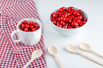 Cornel berries in bowl and cup with wooden spoons high angle view on picnic cloth and white background