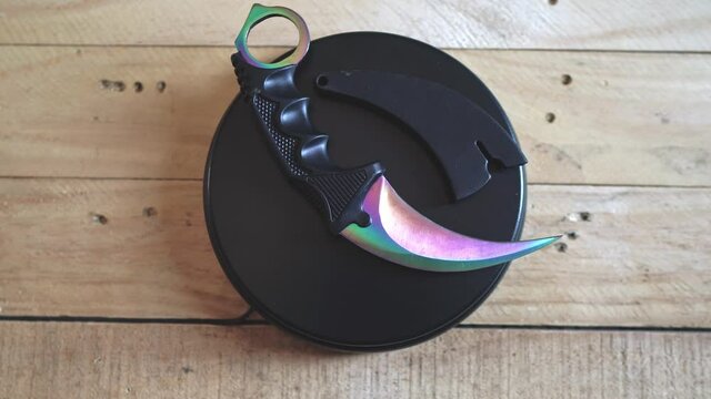 Purple and green karambit knife rotating upon a wooden surface. Reflexive blade.   
