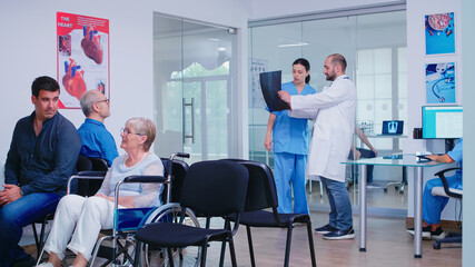 Obraz premium Doctor holding patient x-ray explaining diagnosis to nurse in waiting area. Disabled senior woman wheelchair waiting for medical examination. Hospital and health care system, medicine private clinic