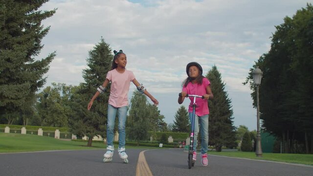 Lovely african american teenage girl roller skating with her younger cute elementary age sister in protective helmet riding on push scooter, enjoying leisure and freedom together in summer park.