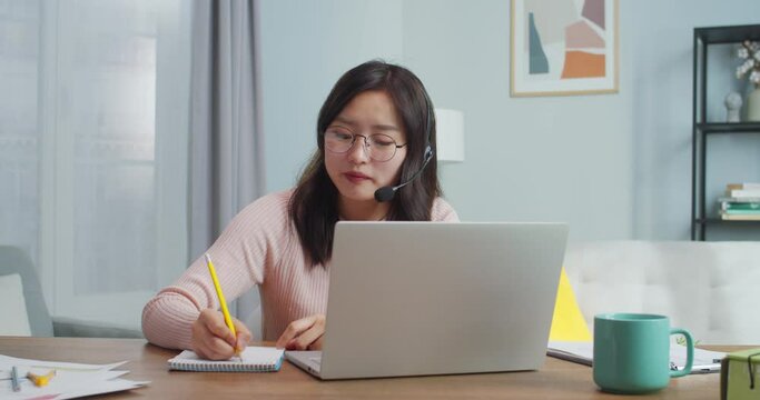 Pretty Asian worker sitting at table and talking to clients, customer via video webcam chat. Hardworking adult woman in headphones and glasses working at home office and taking notes. Support service.