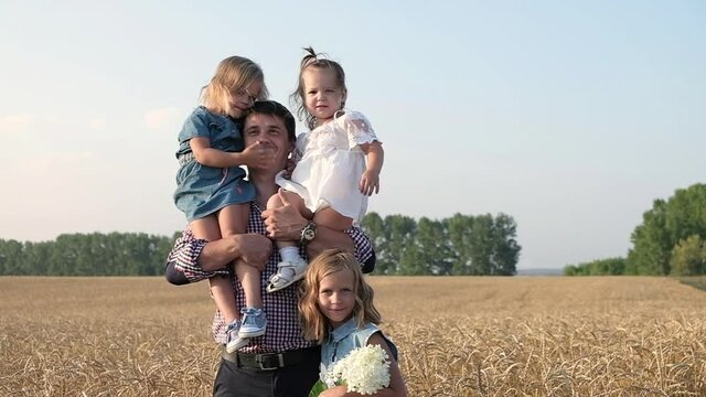 happy father walks through Golden field of wheat with three daughters. man holds girls on shoulders and laughs. Walk through field in summer with family and children. outdoor recreation with parents