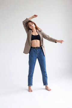 portrait of young caucasian woman with long hair in black top, blue jeans and suit jacket isolated on white studio background. pretty girl posing with her hands raised and bare feet