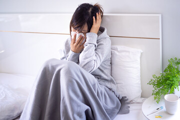 A woman suffering from a headache due to the flu sits wrapped in a blanket on her bed. Woman is wiping off the juice after a high fever. Sick woman concept