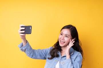 Naklejka premium A cheerful Asian woman making a video call with her lover. Young woman taking a smartphone to take a selfie with the front camera standing on the orange background.