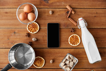 cooking, christmas and seasonal drinks concept - smartphone, ingredients for eggnog, pot on wooden background