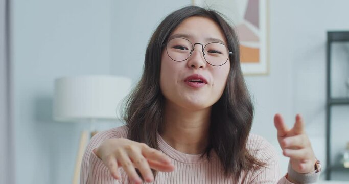 Beautiful Asian woman in glasses sitting at table and talking to camera indoors. Smiling young female vlogger making live stream, video conference fro viewers. Blogging, social media, Internet.