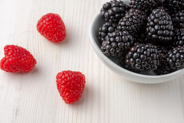 Close-up top view of white bowl with blackberries on white wooden table with raspberries, horizontal, with copy space
