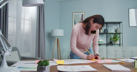 Attracrive adult woman working with charts, diagrams, documents at home. Busy Asian worker talking to client, customer, collegue and taking notes on color stickers. Business, job.