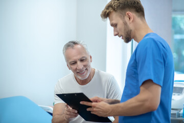 Obraz na płótnie Canvas Young bearded doctor showing clip folder to mature male patient