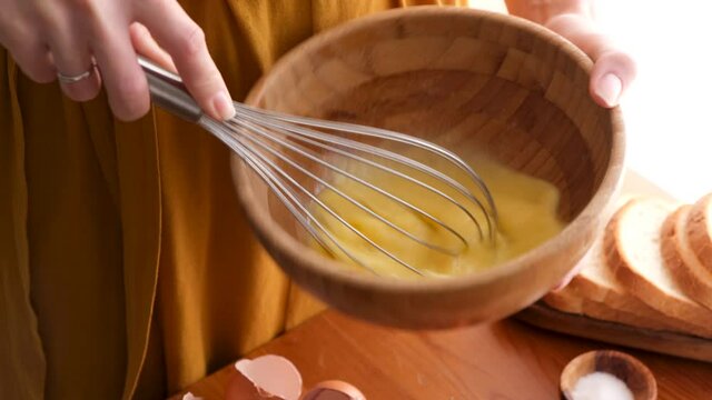 Woman whisking eggs in wooden bowl with metal wire whisk. Cooking eggs, food preparation process in 4K