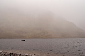 Silhouette of a small fishing motor boat in a lake, Huge mountain covered with fog in the background. Connemara, Ireland.