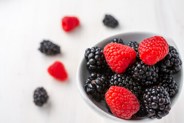 Close-up of white bowl with blackberries and raspberries, on white background with out-of-focus fruits, horizontal, with copy space