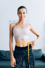 Young sportswoman looking at camera while measuring waist with tape at home