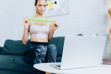 Selective focus of laptop on coffee table and sportswoman holding resistance band on couch