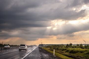 Fototapeta premium Highway after rain. Cars are driving along the road. Grass, trees and bushes grow on the side of the road. Cloudy sky. The sun's rays make their way through the clouds. Fog in the distance.