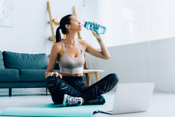 Fototapeta na wymiar Selective focus of sportswoman drinking water while sitting on fitness mat near laptop