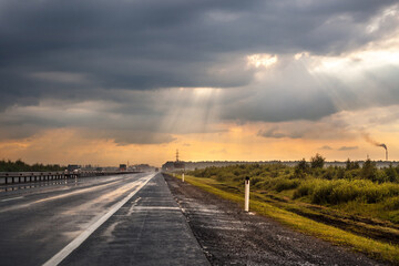 Highway after rain. Cars are driving along the road. Grass, trees and bushes grow on the side of the road. Cloudy sky. The sun's rays make their way through the clouds. Fog in the distance.