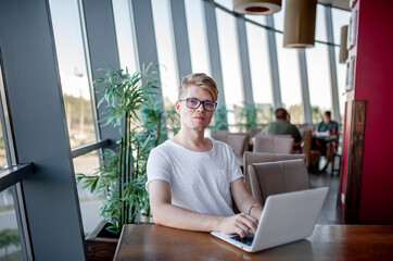 Young guy in a white t-shirt and glasses works on a laptop sitting at a table in a cafeteria