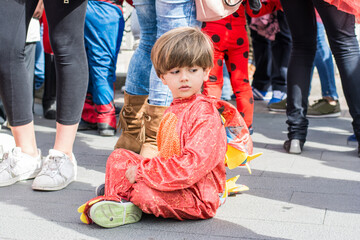 Boy In Dinosaur Costume Sitting On The Ground Surrounded By People.
A little boy prepares for Halloween.