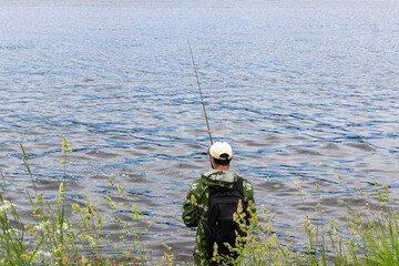 fisherman stands in coastline and fishing on lake or river. Back view of guy holding rod and using it along with fishing line for catching tasty fish in water. International Fishermen's Day concept.