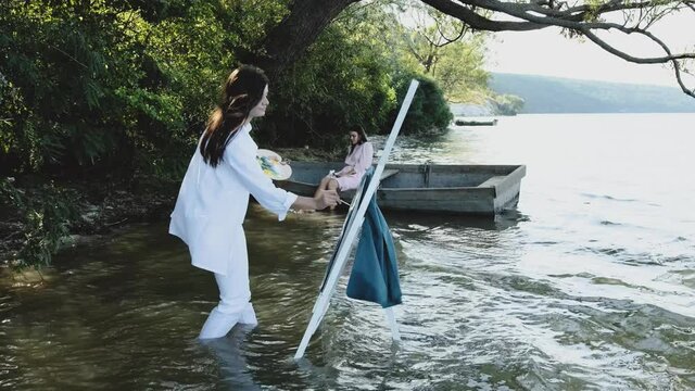 Painter paints on the textile a girl that is sitting in a boat on the shore of a lake. Young woman dressed in a white clothing draws on a jacket.