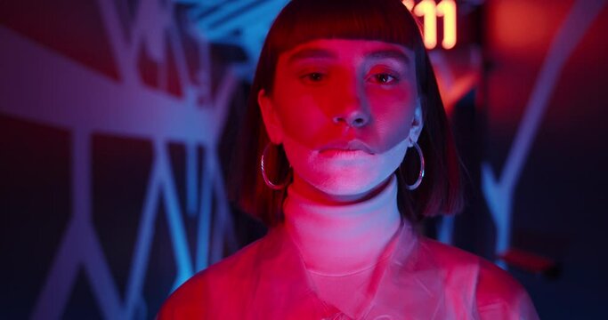Close up view of beautiful woman looking to camera while walking in corridor. Female young person in futuristic outfit and make up. Neon light and glowing numbers at background