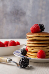 Close-up of plate with pancakes with blackberries and raspberries, fork with blackberries, on white wooden table and bowl with raspberries, gray background, vertical