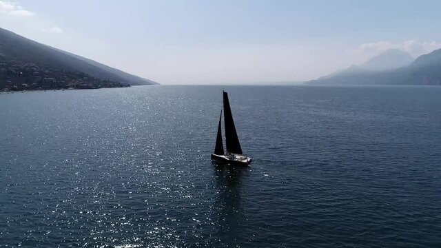 Aerial birds eye view of black sailboat sailing over quiet lake mountainous scenery mountains left and right of the water because of lighting the ship shows as silhouette 4k high resolution footage