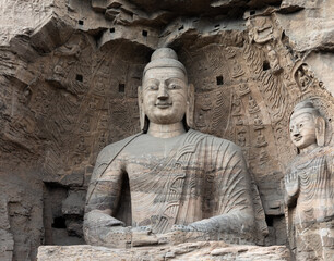 Iconic seated Buddha statue and smaller standing Buddha on right in Cave 20 at Yungang Grottoes, Datong, Shanxi, China. Created by  Monk Tan Yao in 5th century. UNESCO World Heritage.