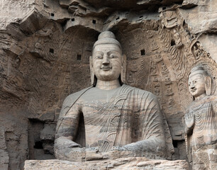 Iconic seated Buddha statue and smaller standing Buddha on right in Cave 20 at Yungang Grottoes,...