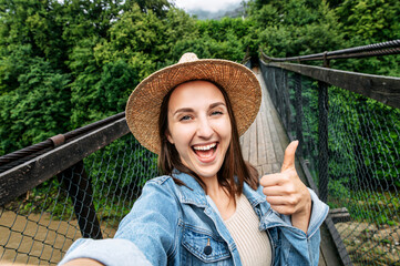 An attractive excited woman in straw hat stands on the suspension bridge, holds a smartphone in...