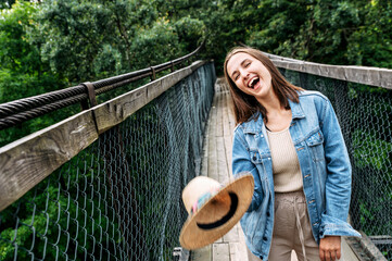 Young cheerful woman stands on suspension cable bridge among wild forest, throws her straw hat and laughing happily. Good mood for adventures